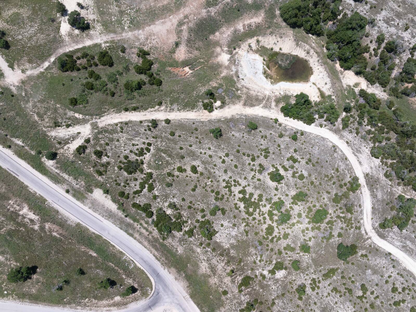 Overhead drone photograph of the 8.3-acre tract as it stands: oak and cedar scrub over pale rocky ground, a caliche ranch track curving across the lot, the stock pond at the top right, and the paved edge of Falling Oak Drive at the lower left.