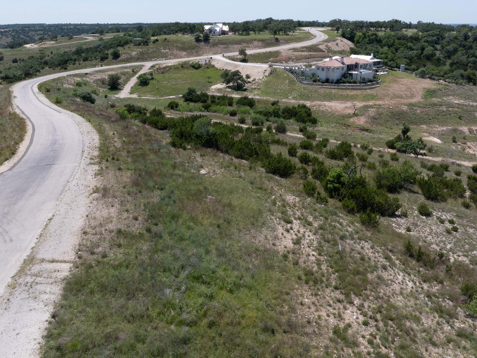 The paved subdivision road curving past the tract's frontage, with two neighbouring custom homes on the rise beyond and open Hill Country running to the horizon.