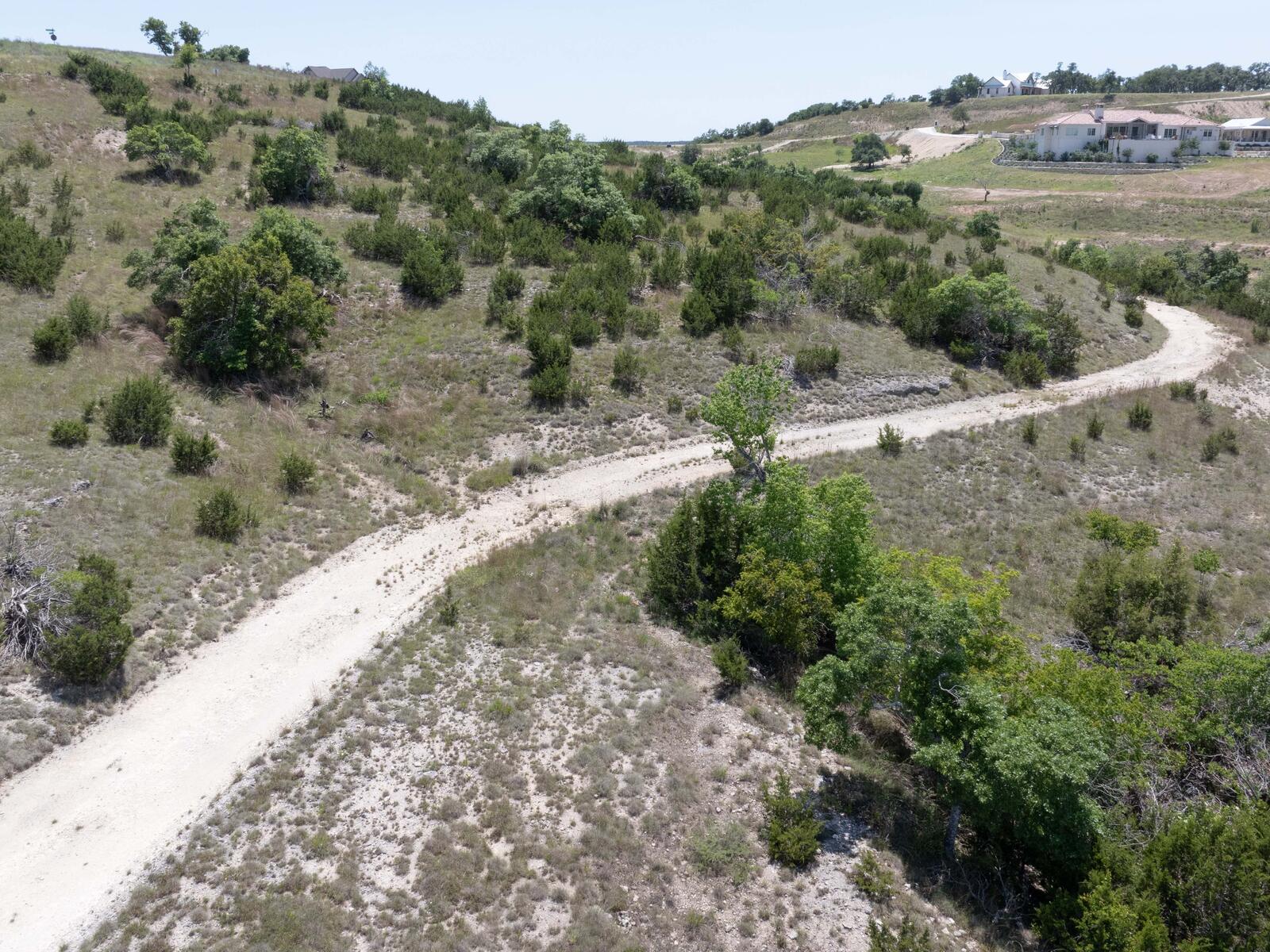 Caliche ranch track curving through the tract's scattered oak and cedar, the ground falling away to the north-east with neighbouring Hill Country homes on the far ridge.