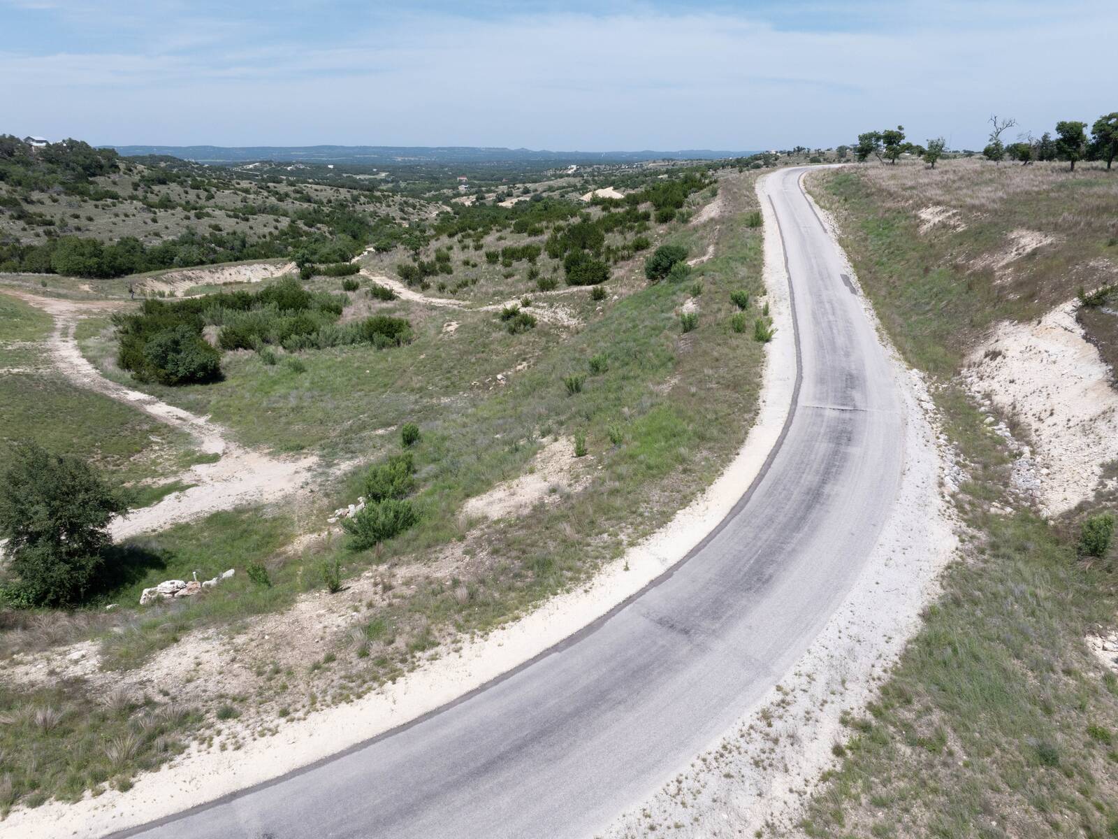 Low-level view along the paved Falling Oak Drive frontage on the lot's south-west edge, the road curving downhill between grassy cut banks with the Hill Country opening out beyond.