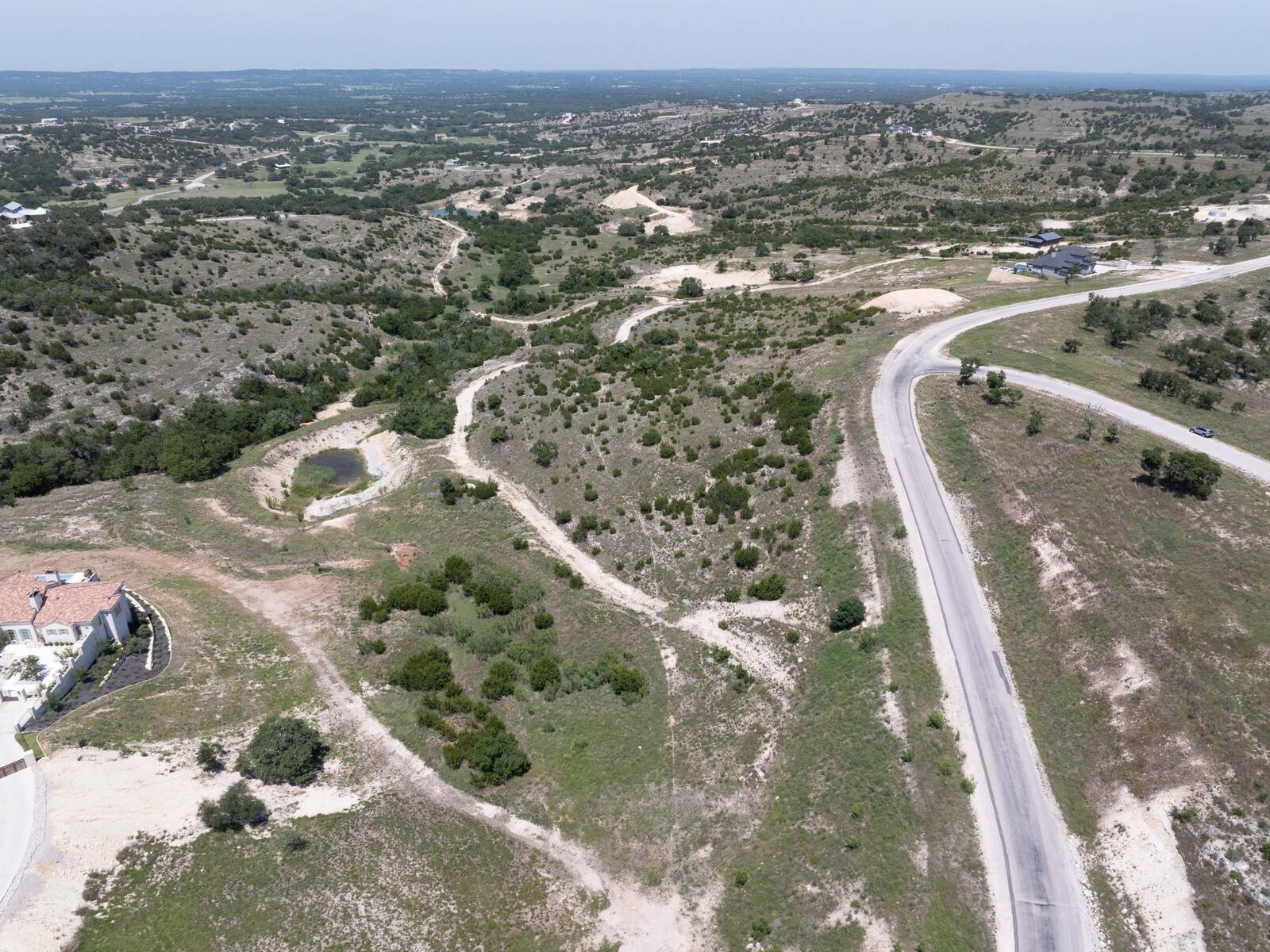 Aerial view across the tract toward the Hill Country horizon, with the stock pond at the centre left, a neighbouring house at the lower left, and the paved Falling Oak Drive frontage running down the right-hand side.